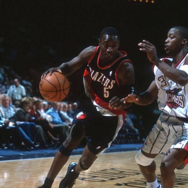 A basketball player in a black Portland Trail Blazers jersey dribbles past a defender in a white Houston Rockets uniform during a game, capturing the fast-paced energy reminiscent of the golden age of basketball on a brightly lit court with fans watching.