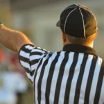 A football referee is seen from behind, wearing a black and white striped shirt and black cap, raising his left arm and pointing left, likely signaling a call on the field in accordance with national football league rules, with blurred players and background.
