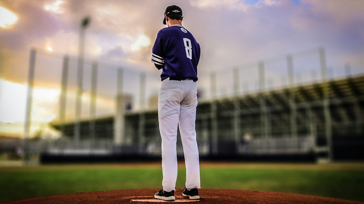 Pitcher in a purple jersey standing on the mound at dusk, seen from behind