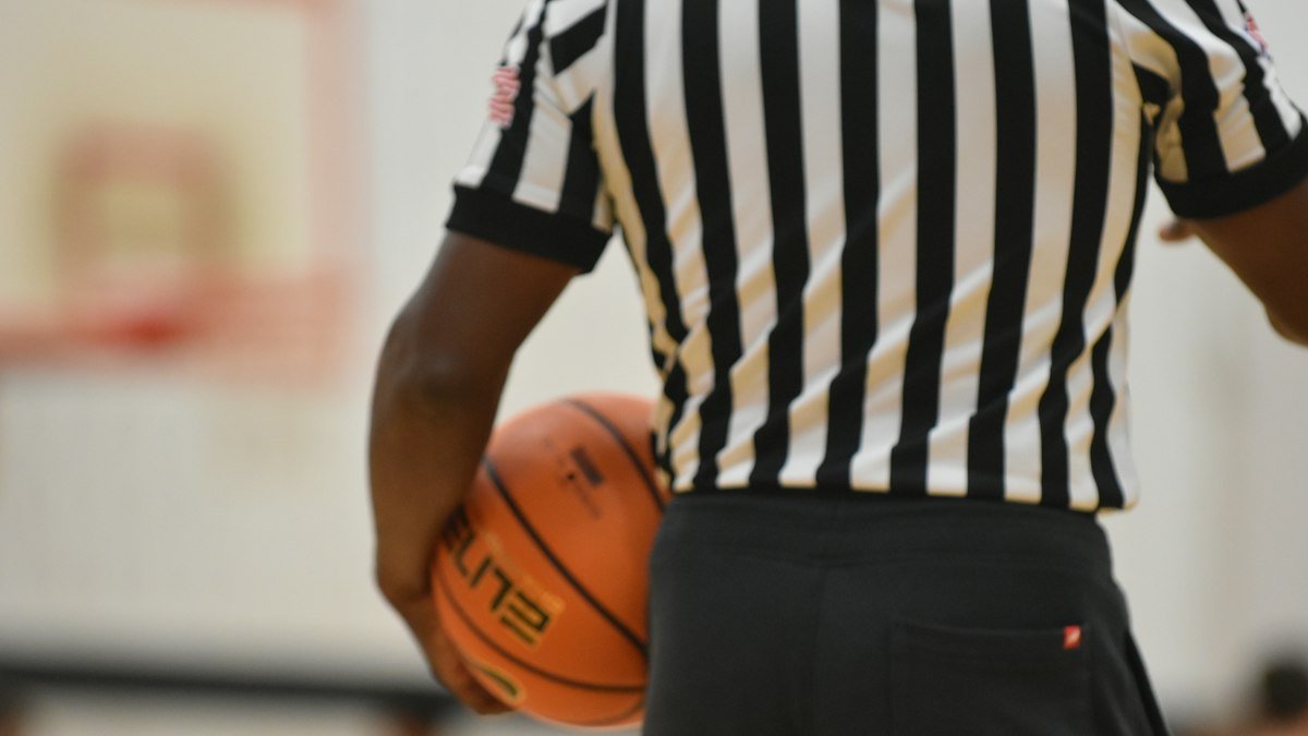 A basketball referee in a striped shirt holds a ball against his hip on a gym floor