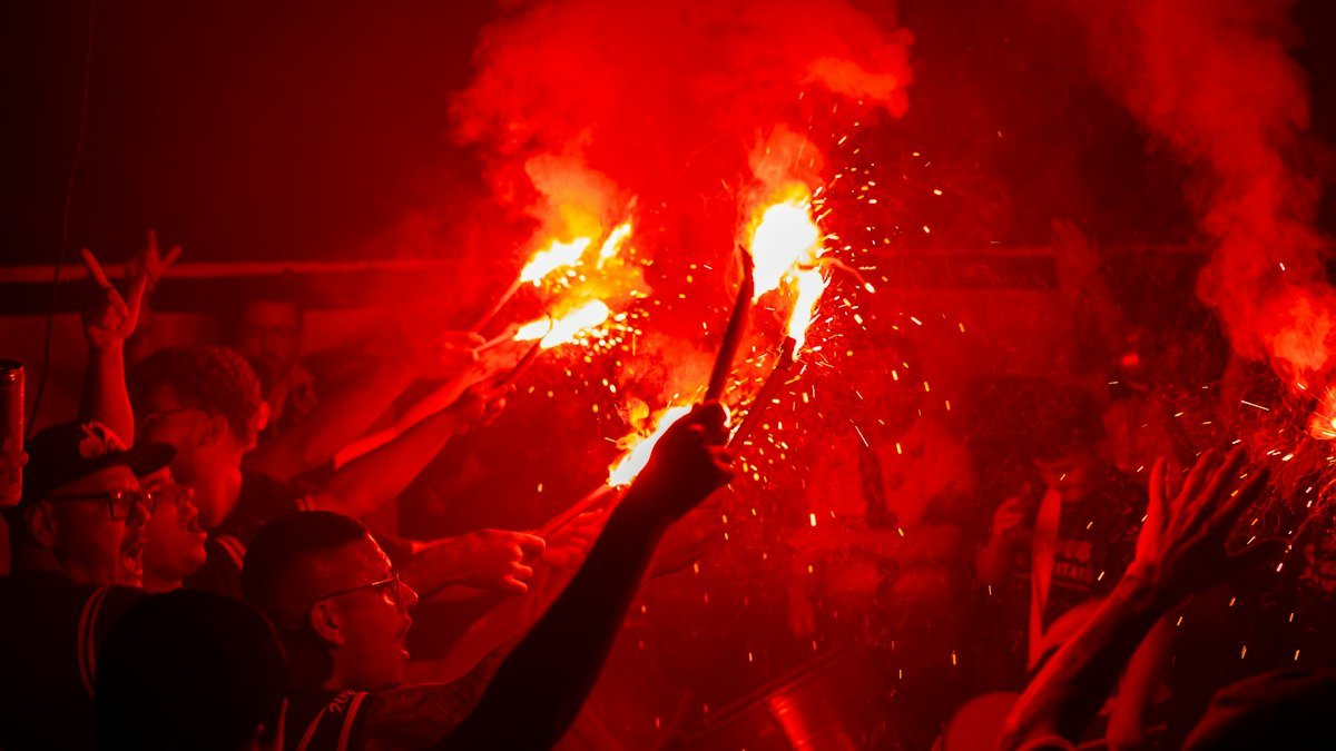 Supporters at night lighting red flares in a stadium crowd, with smoke and sparks rising