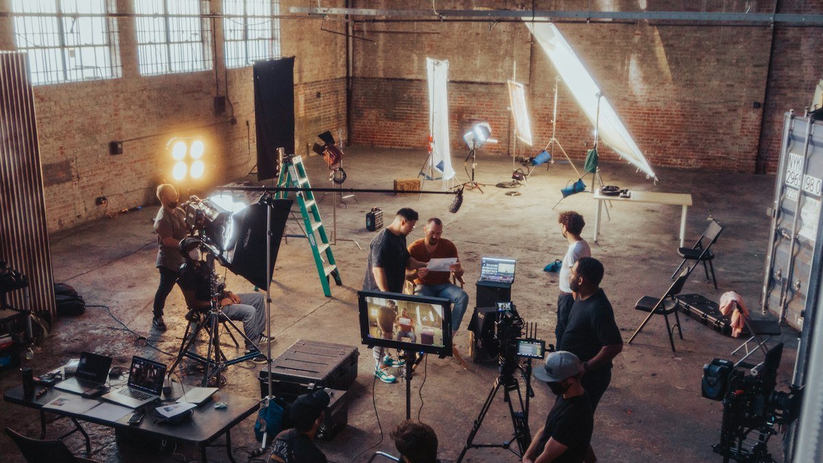 A film crew gathers around a camera and monitors under studio lights in a large brick warehouse