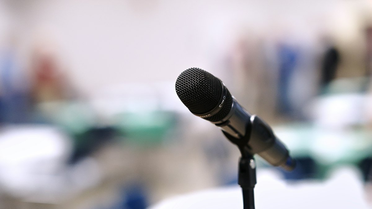 A black microphone on a stand with a blurred room of tables and people behind it