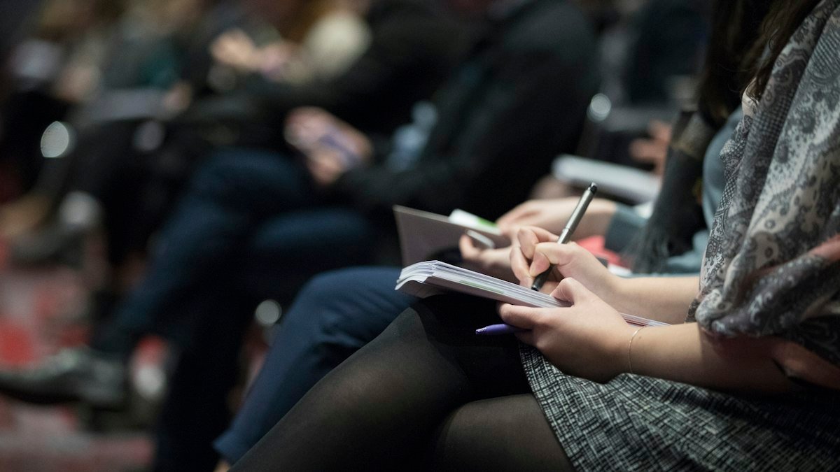 People seated in a row writing in notebooks at an event, with the rest of the audience blurred behind them