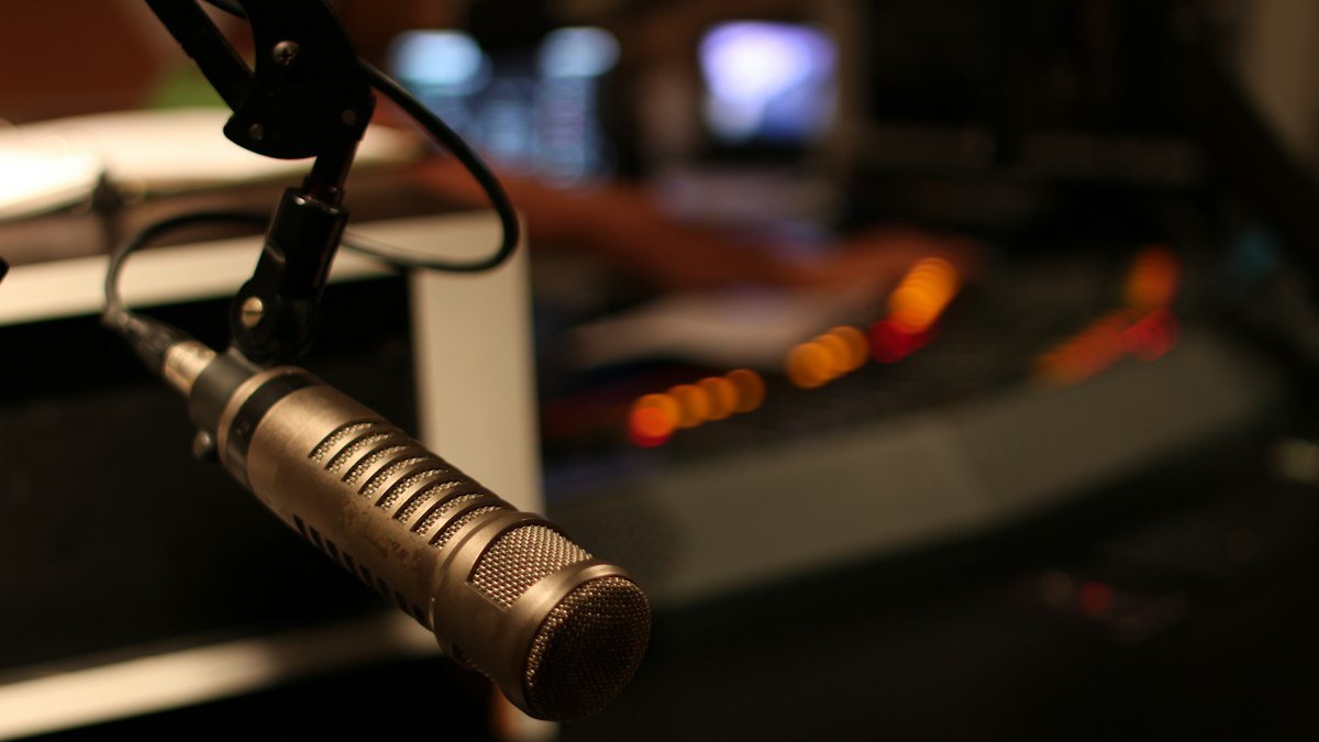 Close-up of a studio microphone on a boom arm in front of a blurred mixing desk