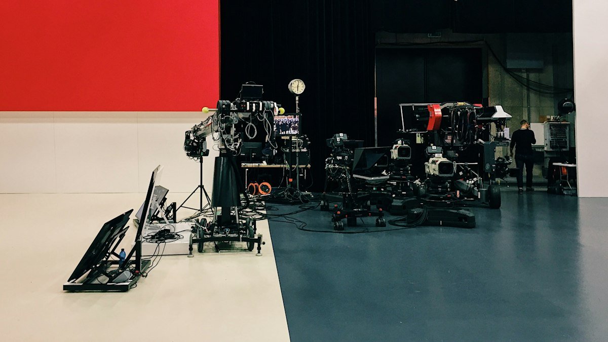 A television studio floor with broadcast cameras and monitors in front of a red and white wall