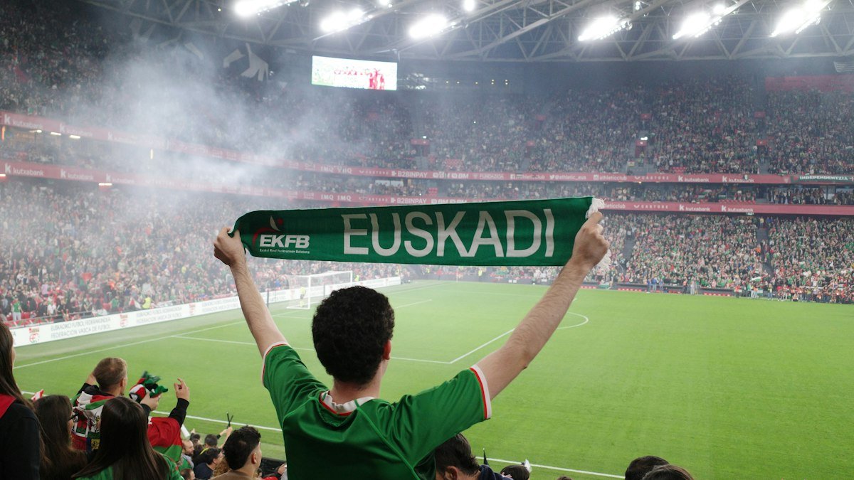 Fan seen from behind holding a green Euskadi scarf above his head in a packed, smoky stadium