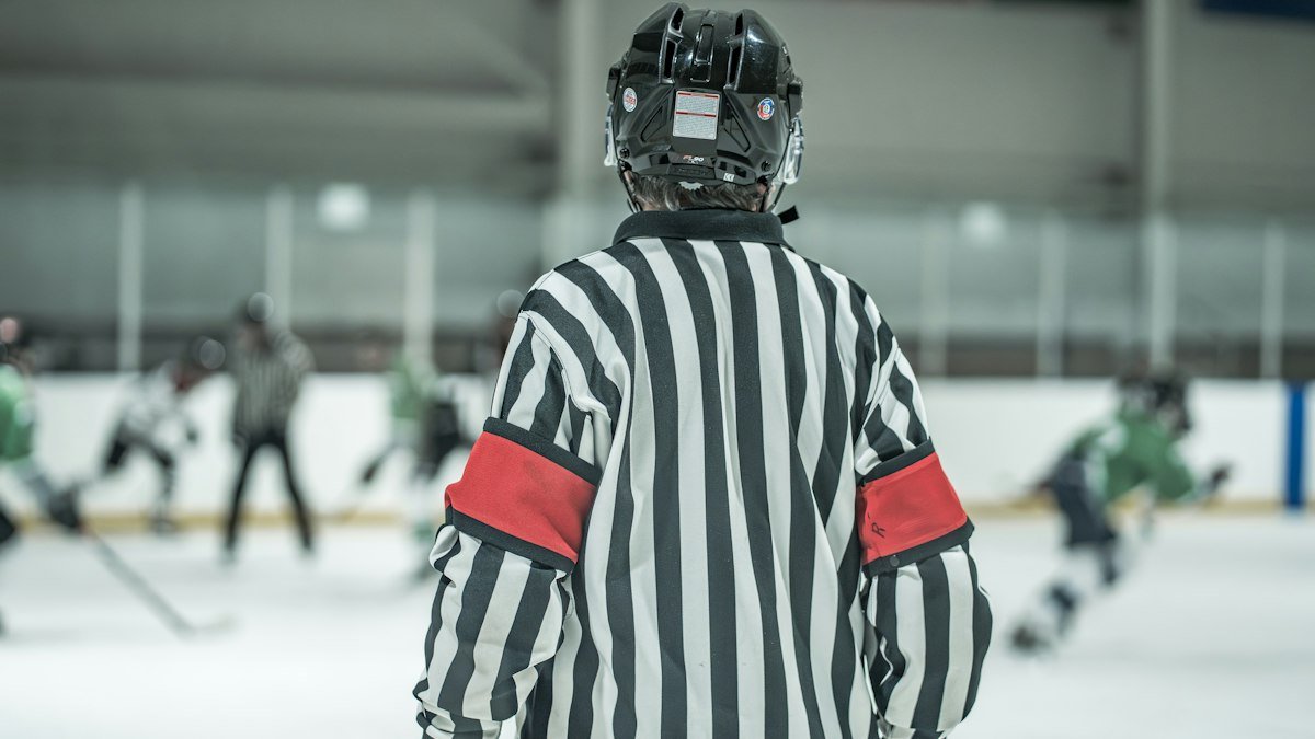 Hockey referee in a black-and-white striped jersey and helmet, seen from behind, watching players on the ice