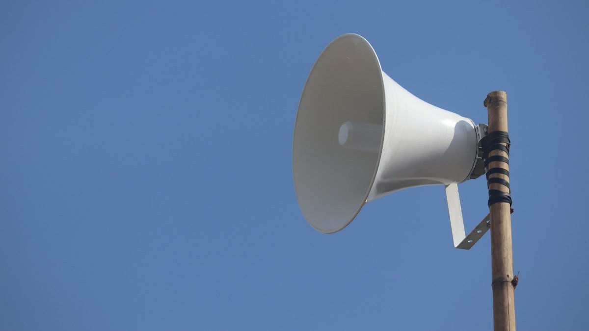 White loudspeaker horn mounted on a wooden pole against a clear blue sky
