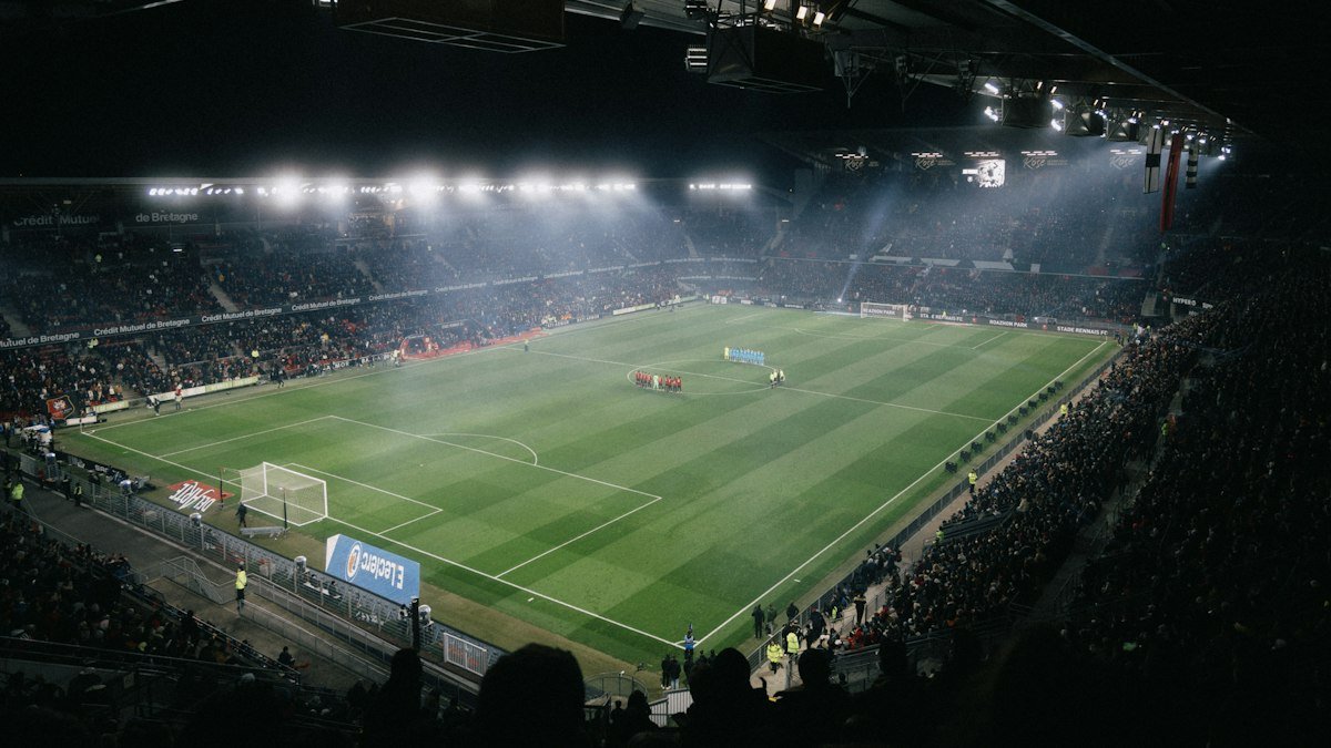 Soccer stadium lit by floodlights at night, with crowded stands around a green pitch