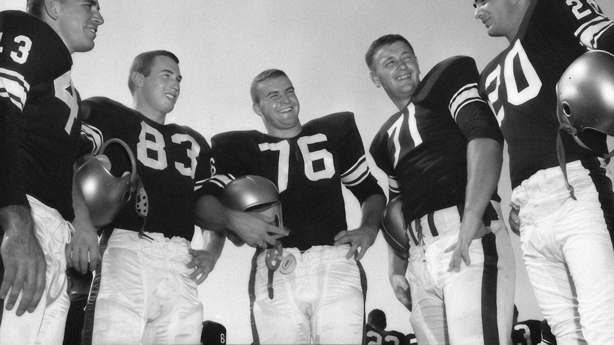 Black-and-white archive photo of five smiling football players in dark jerseys standing together, several holding their helmets